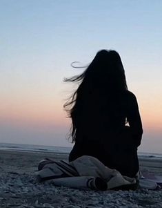 a woman sitting on top of a beach next to the ocean with her hair blowing in the wind