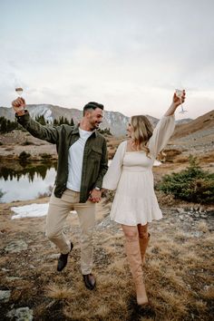 a man and woman holding wine glasses in their hands while walking through the grass with mountains in the background