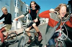 three women riding bikes down the street with their arms around each other's shoulders