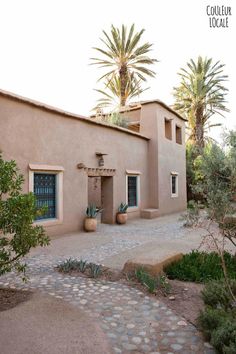 an adobe style house with palm trees in the back ground and cobblestone path leading to it