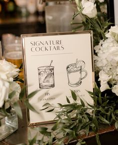 a sign that is on top of a table next to some flowers and glasses with drinks in them