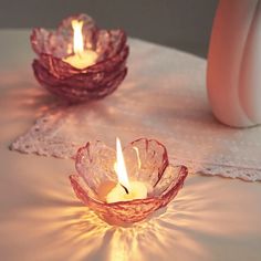 two glass bowls with lit candles in them on a white table cloth next to a pink vase