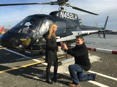 a man kneeling down next to a woman in front of a helicopter on the water