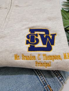 Southwest DeKalb High School Logo Shirt Show your Panther pride with our custom-embroidered Southwest DeKalb High School Logo Shirt! Featuring the iconic SWD logo in bold blue and gold, this shirt is perfect for students, alumni, and supporters alike. The logo is meticulously embroidered on the left chest, ensuring a polished and durable finish that stands the test of time. Make it uniquely yours by adding your name underneath the logo for a personalized touch. Whether you're heading to class, c Cotton Tops With Embroidered Logo In Team Spirit Style, Gold Cotton Crew Neck Shirt, Collegiate Cotton Tops With Embroidered Text, Collegiate Style Cotton Top With Embroidered Text, Collegiate Embroidered Cotton T-shirt, School Spirit Cotton Top With Custom Embroidery, Cotton Tops With Custom Embroidery For School Spirit, Panther Pride, School Pride