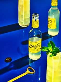 three bottles and two glasses filled with lemonade on a blue counter top next to a gold handled bottle opener