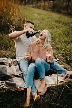 a man and woman sitting on a blanket drinking from wine bottles in the grass with trees in the background