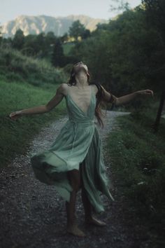 a woman in a green dress walking down a dirt road with her arms spread out