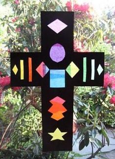 a cross made out of black paper with different colored shapes and sizes on it in front of some flowers