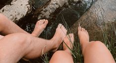 two people sitting next to each other on the ground near water and rocks with grass growing in front of them