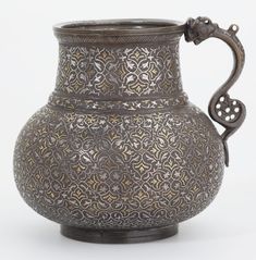 an ornate metal vase with a handle on the top and handles to the bottom, sitting on a white surface