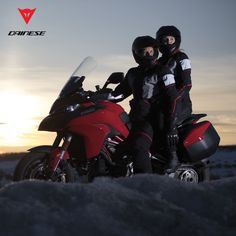 two people are sitting on a red motorcycle in the snow at sunset, with their arms around each other