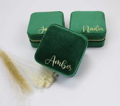 two personalized green velvet jewelry boxes with gold lettering on the front and side, sitting next to a feather