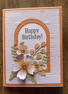 a happy birthday card with flowers and an arch on the front, sitting on a wooden table