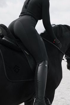 a woman riding on the back of a black horse next to the ocean in wetsuits