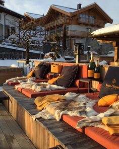 an outdoor lounge area with several pillows and blankets on it, in front of snow covered buildings