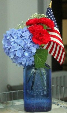 a blue vase with red, white and blue flowers in it next to an american flag