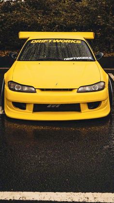 a yellow sports car parked in a parking lot on a rainy day with trees and bushes behind it