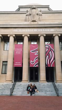 three people sitting on the steps in front of a building with columns and banners hanging from it