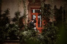 a bride and groom kissing in front of a red door surrounded by greenery at their wedding