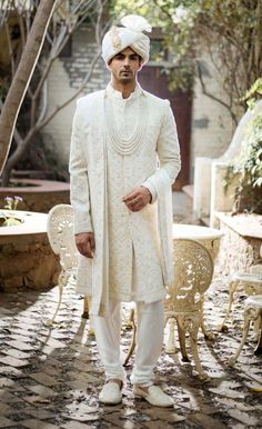 a man in a white outfit standing on a cobblestone walkway with trees and chairs behind him