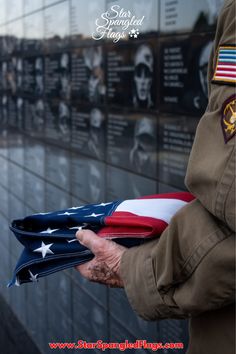 A veteran holding a folded American flag stands solemnly in front of the Vietnam War Memorial.