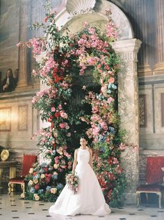 a woman in a wedding dress standing next to a flower covered archway with pink and red flowers