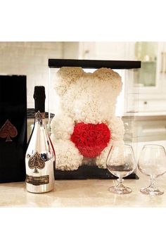 a white teddy bear sitting next to two wine glasses and a bottle with roses in it