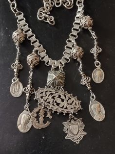 "A bib style assemblage of Victorian silver and religious medals. The central pendant is a Victorian brooch with chased and pierced floral and vine motif. Suspended from the brooch is a Victorian silver stick pin charm monogrammed letter \"C\". The other charm is a hallmarked sterling Victorian watch fob with the initials \"GW\" inside a heart. On the back is in engraved \"AL to GW \" and dated \"24/3/04\" for March 24, 1904. I've used a vintage silver plated Art Deco style bracelet link to attach it to the silver plated book chain.  The accompanying pendants are Bali sterling beads, faceted pyrite and religious Catholic medals from the 1930s or older featuring Our Lady of Lourdes. I believe they are plated silver. Finishes with larger links and hook so length is adjustable." Victorian Metal Medallion Jewelry, Antique Silver Jewelry With Large Pendant, Victorian Style Metal Medallion Jewelry, Ornate Pendant Necklace With Charms, Ornate Pendant Necklaces With Charms, Ornate Vintage Charm Pendant Jewelry, Antique Silver Pendant Jewelry With Charms, Ornate Pendant Jewelry With Vintage Charm, Silver Medallion Necklace With Charms