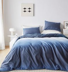 a bed with blue sheets and pillows in a white room next to a painting on the wall