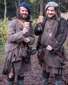 two men dressed in period clothing holding coffee cups and walking through the woods on a trail