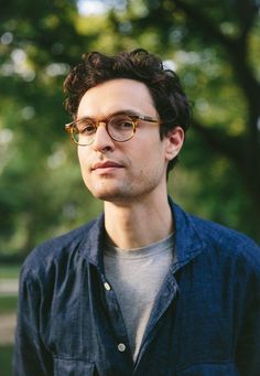 a young man wearing glasses standing in front of trees and looking off into the distance