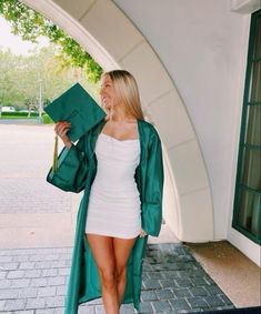 a woman in a short white dress and green coat holding a graduation cap while standing on the sidewalk