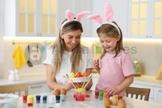 two girls in bunny ears painting easter eggs with colored watercolors on the table