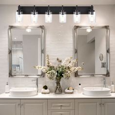 two sinks and mirrors in a bathroom with lights on above them, along with vases filled with flowers