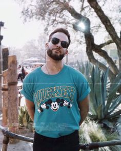 a man standing in front of a cactus with sunglasses on his face and wearing mickey mouse t - shirt