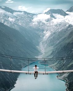 a man standing on a bridge over a river in the middle of mountains with snow capped peaks behind him