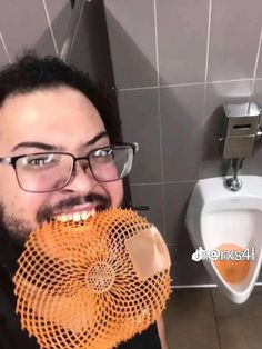 a man with glasses holding an orange object in front of his face while standing next to a urinal