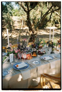 an outdoor table set with plates and place settings for dinner under the shade of a tree