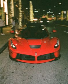 a red sports car is parked on the side of the road at night with people walking by