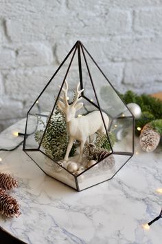 a glass terrarium with a deer and pine cones in it on a marble table