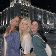 three young women standing in front of a large building at night with the lights on