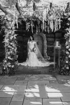 a woman in a wedding dress standing under an archway with flowers on the wall and greenery