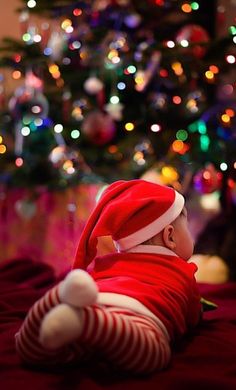 a small baby in a santa hat laying on a bed next to a christmas tree