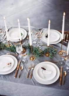 the table is set with white plates and silverware, gold candlesticks, greenery, and napkins