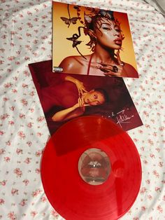 two red vinyl records sitting on top of a bed next to an album with the same cover