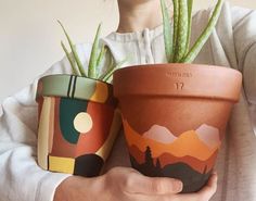 two potted plants with mountains and trees painted on them, one is holding the other