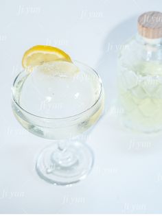 a glass filled with ice and lemon on top of a white table next to a bottle