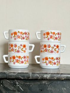 three white cups with orange and pink flowers on them sitting on a marble countertop