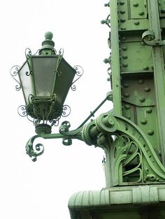 an old fashioned street light on top of a green building with ornate ironwork around it