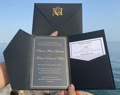 a person holding up an open black and gold wedding card with the ocean in the background